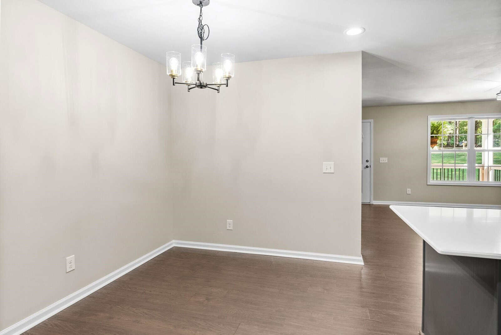 Empty residential dining area with hardwood flooring, bright walls, and a modern chandelier, adjacent to a kitchen with white countertops.