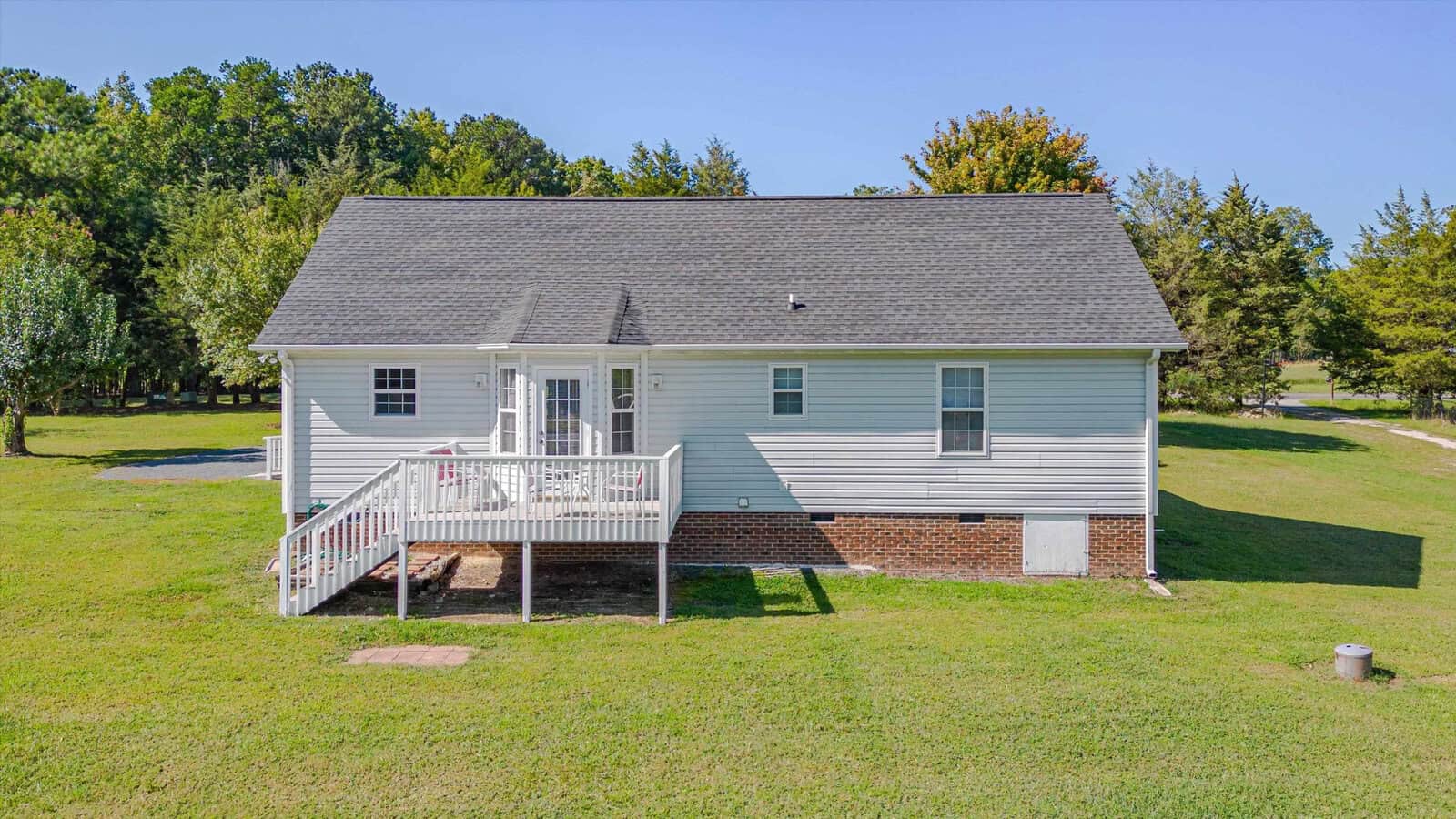 A small white house with a gray shingle roof, a wooden back deck with stairs, and a brick foundation, surrounded by a large grassy yard and trees under a clear blue sky.