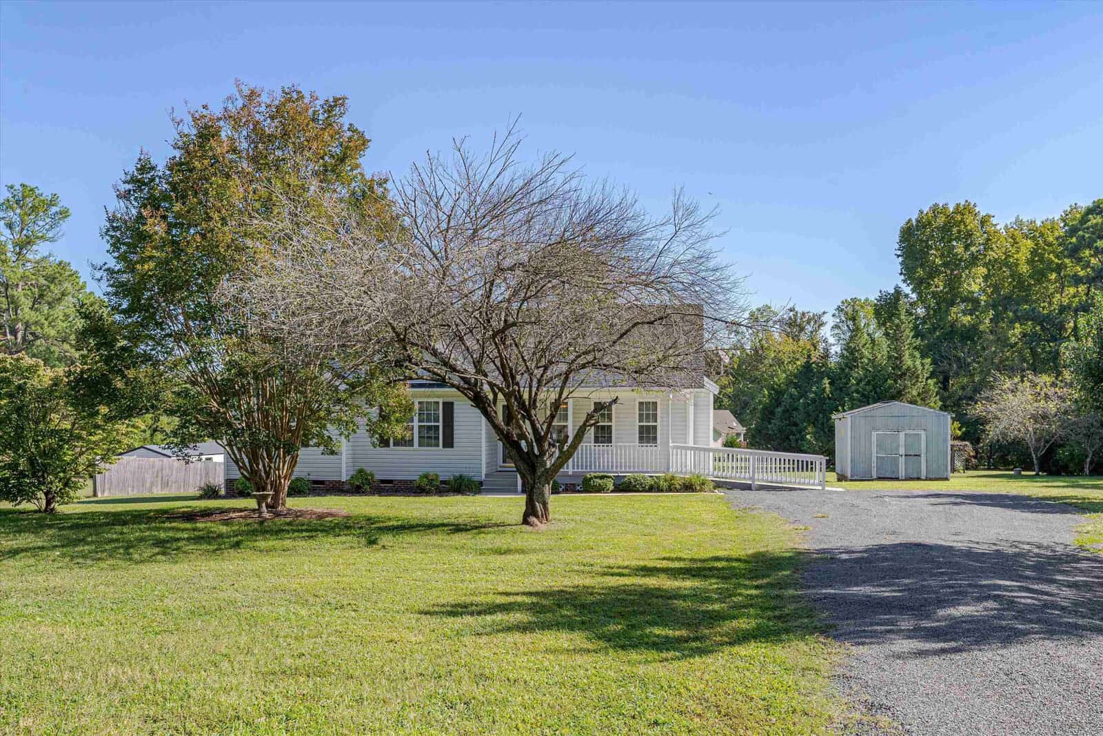 A white house with a front porch and ramp sits behind a large yard with trees. A gravel driveway leads to a white shed in the background. The sky is clear and blue.
