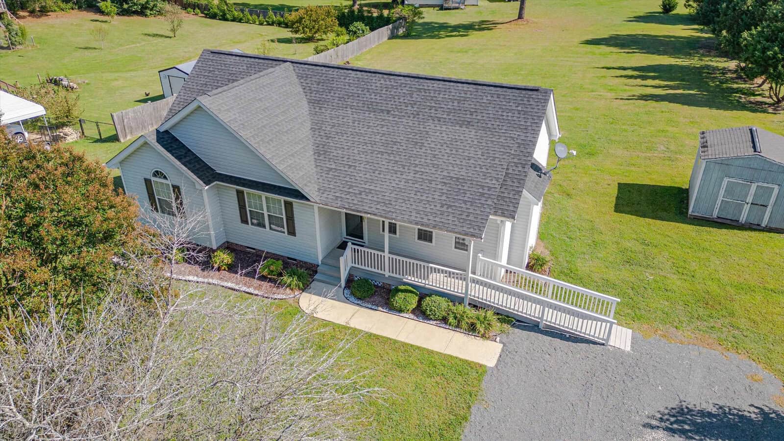 Aerial view of a small, single-story house with gray siding and a covered front porch, surrounded by a well-kept lawn, bushes, a gravel driveway, and a storage shed in the backyard.