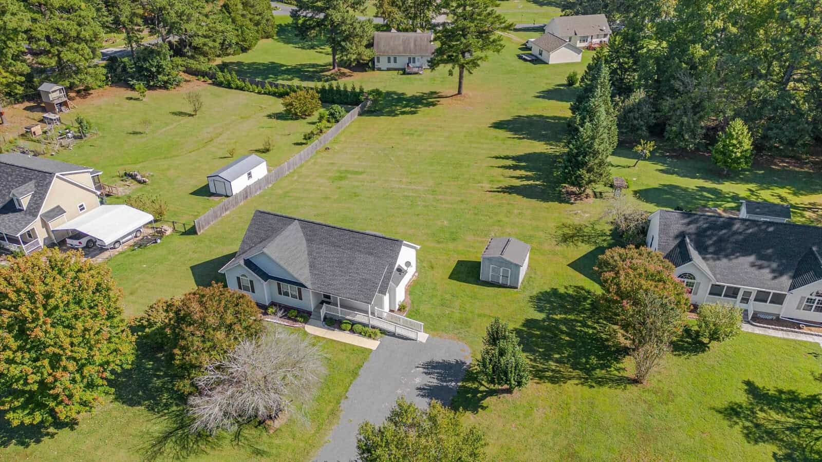 Aerial view of a suburban neighborhood with green lawns, several houses, garden sheds, trees, and a driveway leading to a white house in the center of the image.