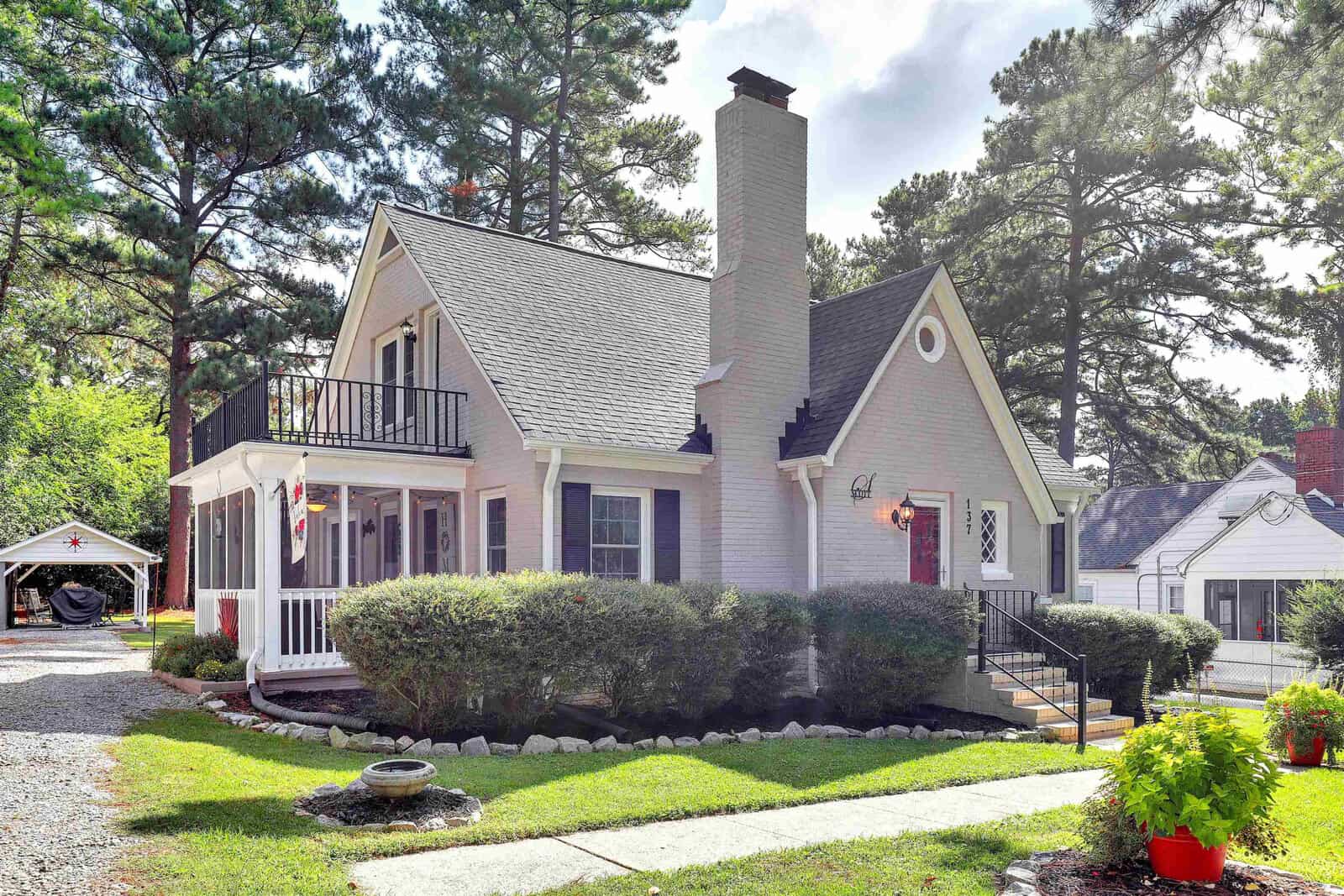 A cozy, two-story house with a white exterior and gray roof, featuring a chimney and a small front porch, surrounded by greenery and a well-manicured lawn, with a driveway leading to a carport on the left side.