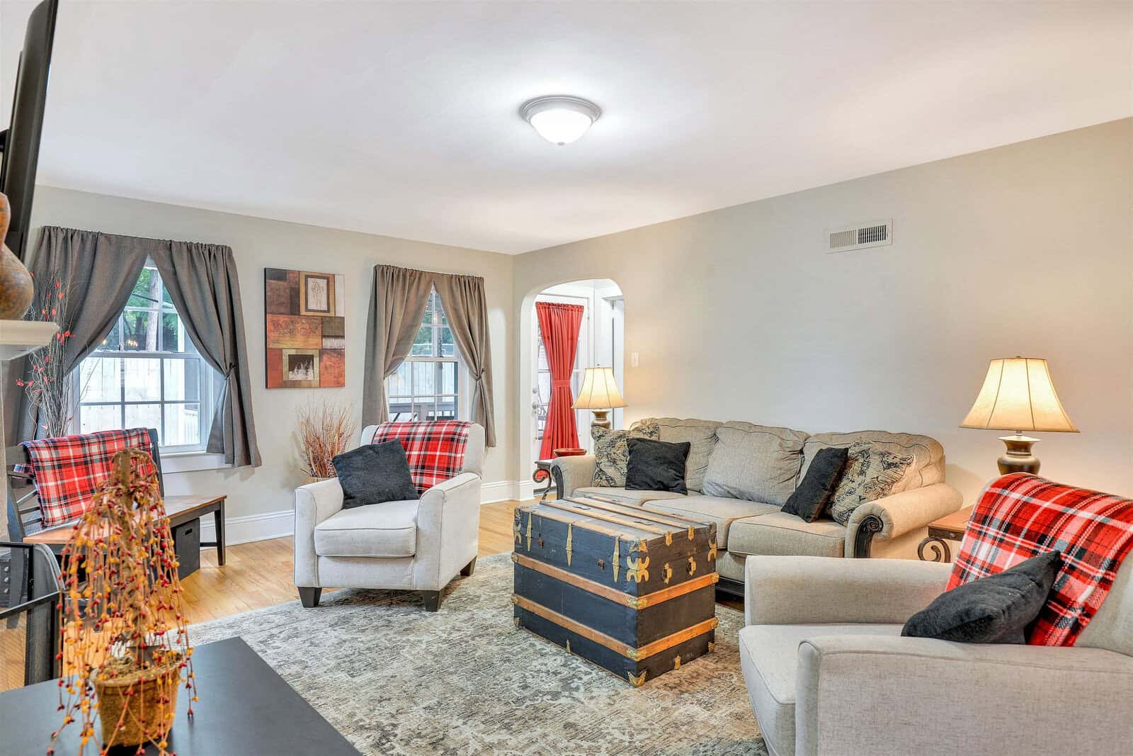 A cozy living room with neutral walls featuring a grey sofa, two matching armchairs with red plaid throws, a central wooden chest used as a coffee table, and decorative lamps on side tables. the room is accented by red curtains, a beige area rug, and wall art, creating a warm and inviting space.