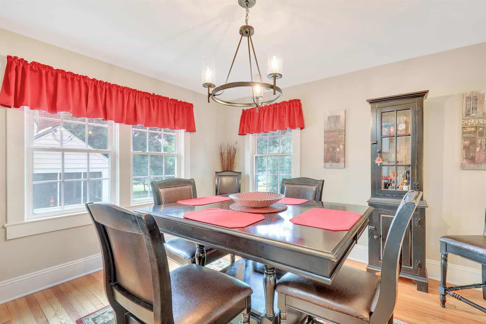 A well-lit dining room featuring a wooden table set with red placemats, surrounded by six chairs, red window curtains, and a hanging chandelier, with a view looking out to the window and hardwood floors.