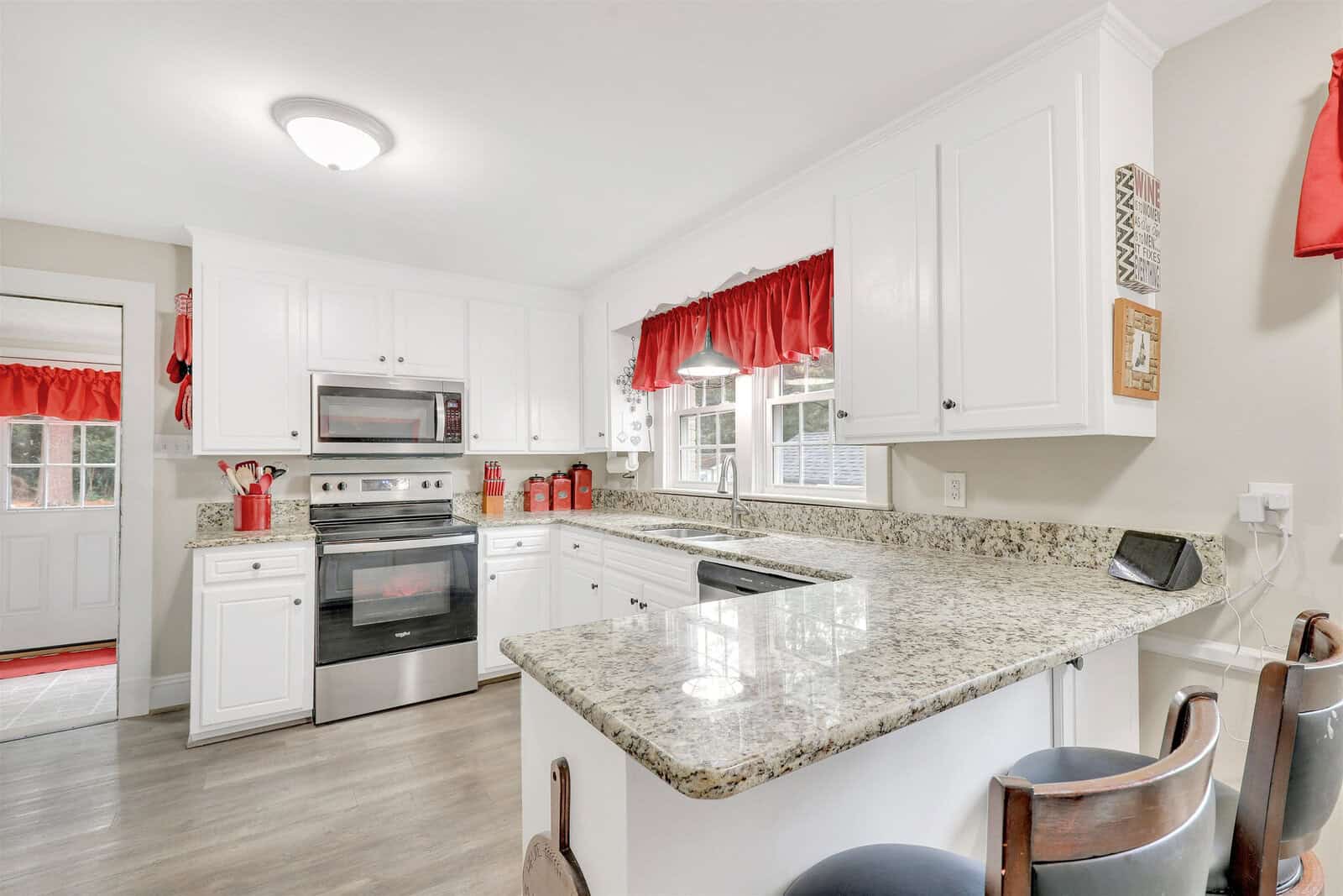 A clean, modern kitchen with white cabinetry, granite countertops, stainless steel appliances, and red window treatments.