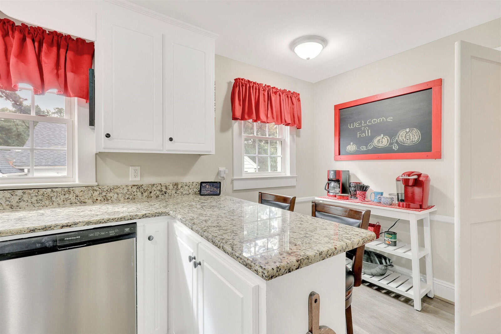 A kitchen interior featuring white cabinetry, granite countertops, and red accents including window curtains, kettle, and decorations, with a chalkboard sign welcoming the fall season.