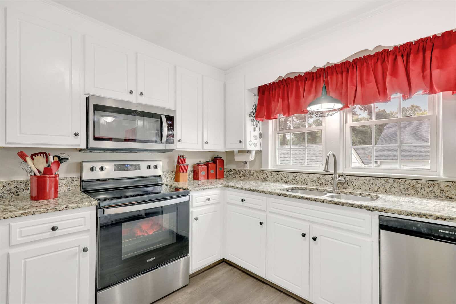 A clean, modern kitchen with white cabinetry, stainless steel appliances, granite countertops, and red window valances.