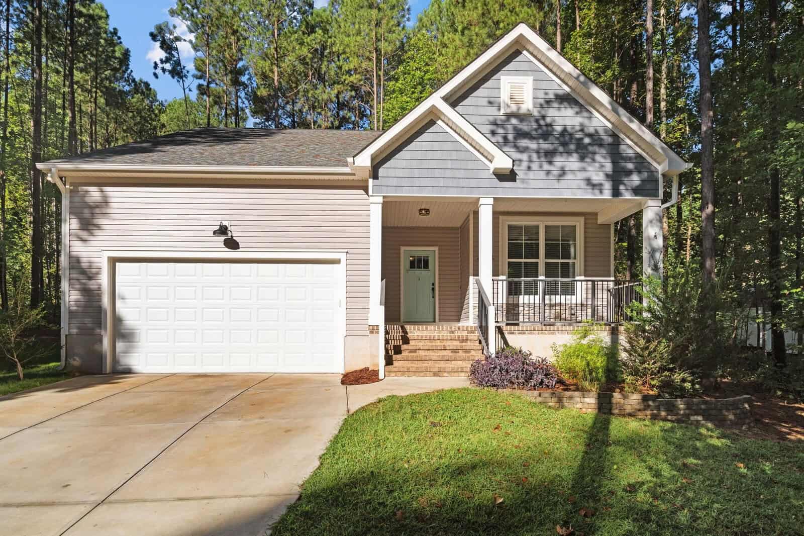 A two-story house with a gray and white exterior is surrounded by tall trees. It has a two-car garage, a small covered porch, and a well-maintained lawn. The driveway is concrete, and there are steps leading to the front door.