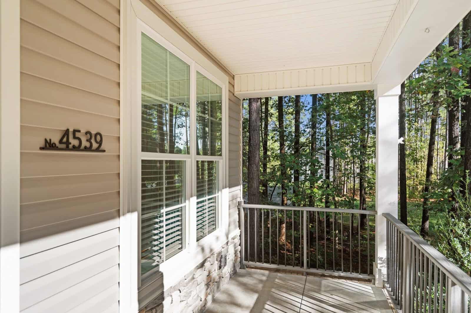 A front porch with a beige siding exterior and a house number "4539" displayed. The porch has white railings and overlooks a wooded area with tall trees. Sunlight filters through the trees, casting shadows on the ground.