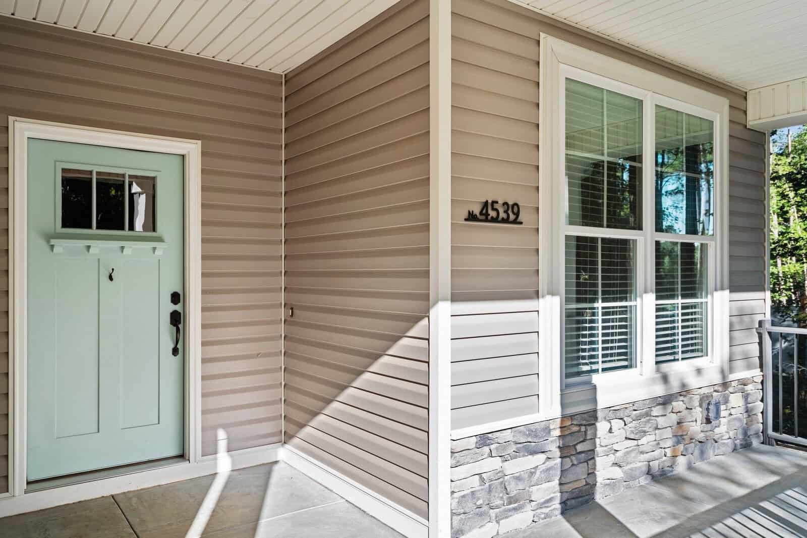 Front porch of a house with beige siding and stone accents. A light teal door with a small window, next to a large, multi-paned window with white trim. House number 4539 is mounted beside the window. The porch has a light gray floor.