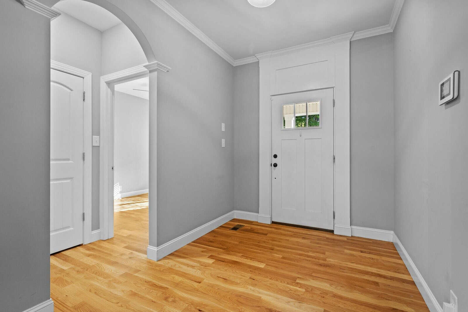 A small, bright entryway features light gray walls, a white door with a window, and hardwood flooring. An arched doorway and a white closed door are visible on the left. Natural light streams from a room to the side.