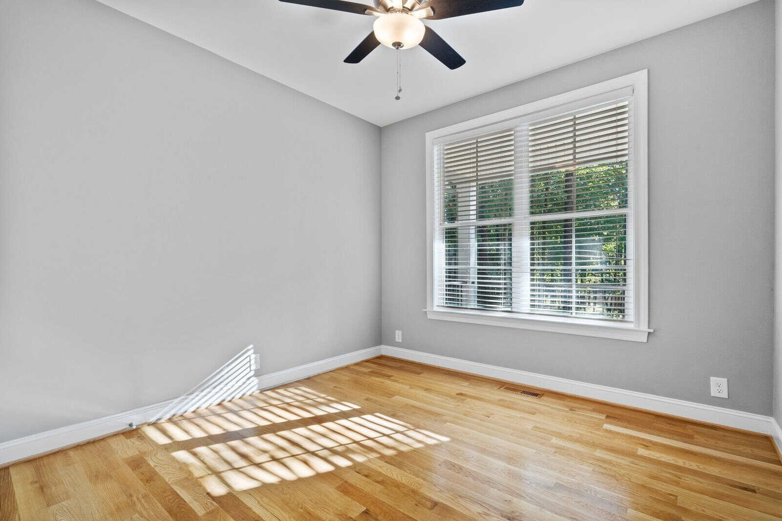An empty room with light gray walls and a wooden floor. A ceiling fan with a light is on the white ceiling. Sunlight filters through a large window with blinds, casting shadows on the floor.