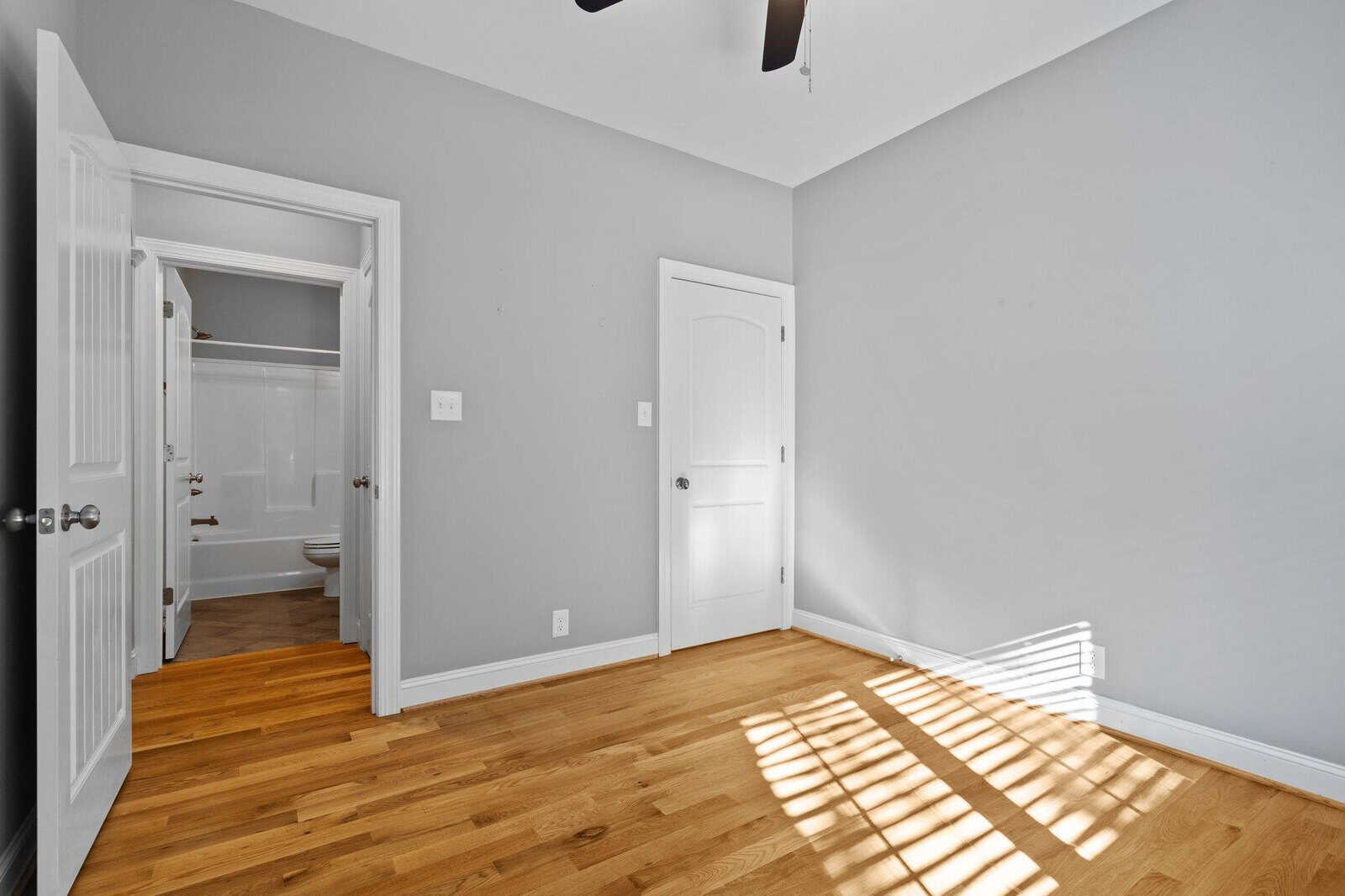 A bright, empty room with light wooden flooring and gray walls. There is a white door leading to a bathroom with visible white fixtures. Sunlight streams through windows, casting shadows on the floor. A ceiling fan is partially visible above.