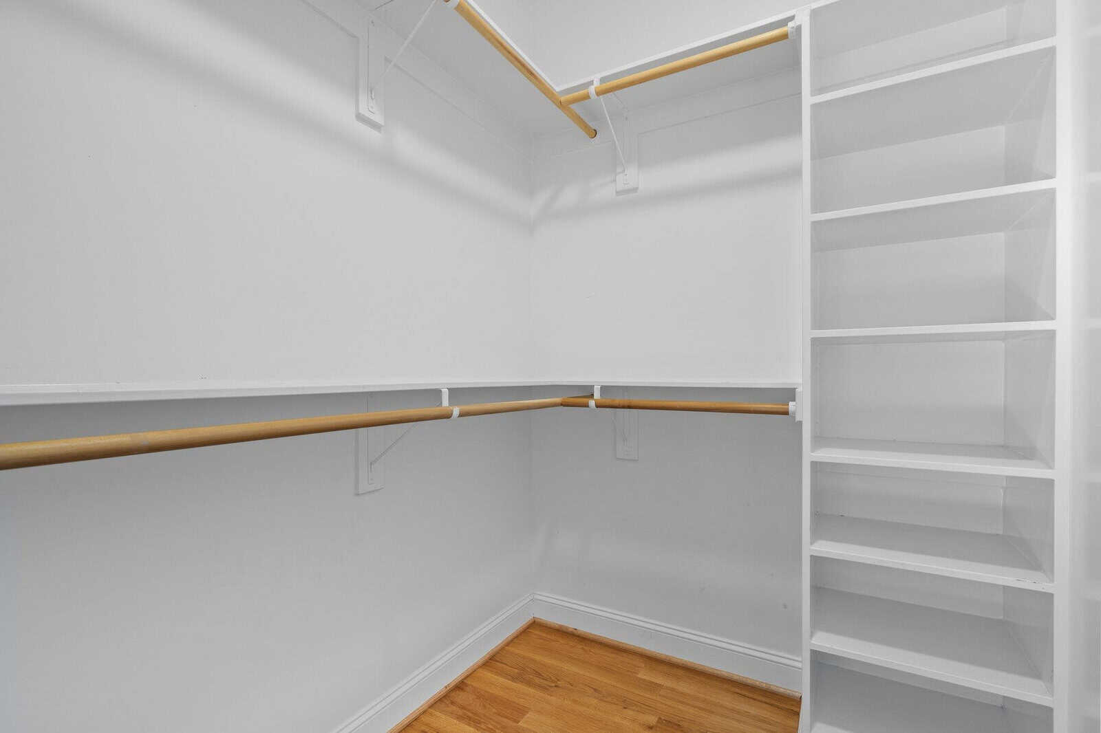 A white, empty walk-in closet features wooden rods for hanging clothes and built-in shelves on the right side. The floor is made of polished wood.
