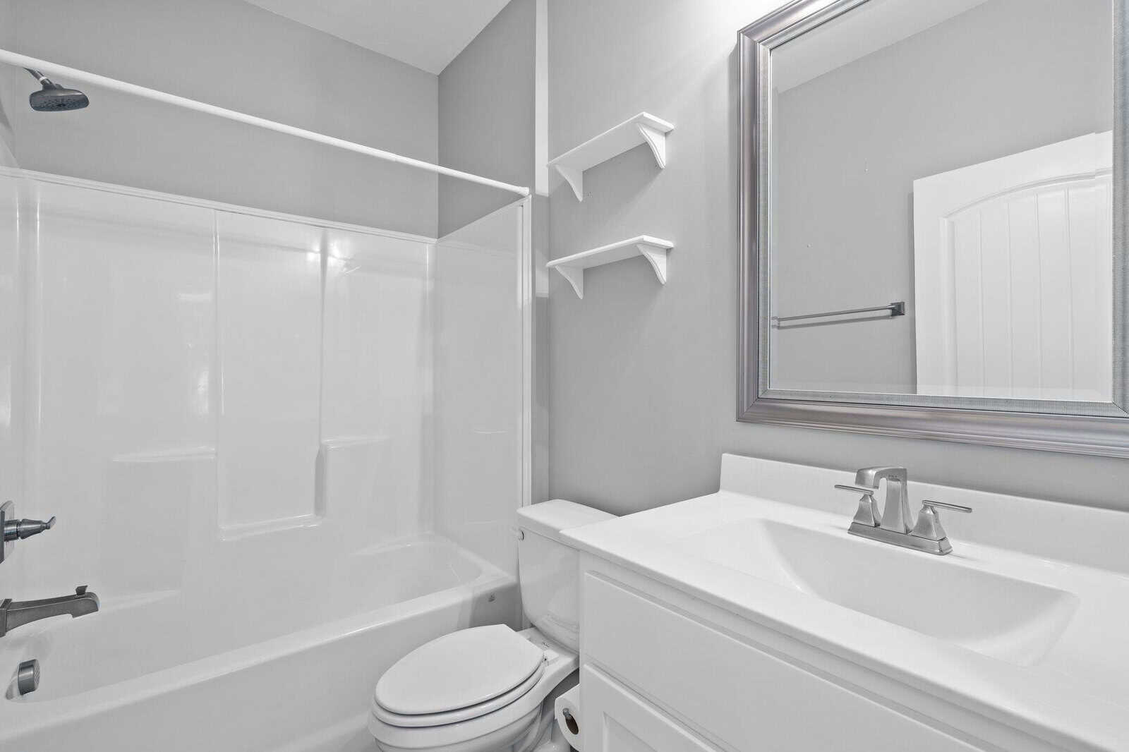 A modern bathroom with white walls and fixtures, featuring a bathtub with a showerhead, a toilet, a white countertop with a silver faucet, a large rectangular mirror, and two small white shelves on the wall.
