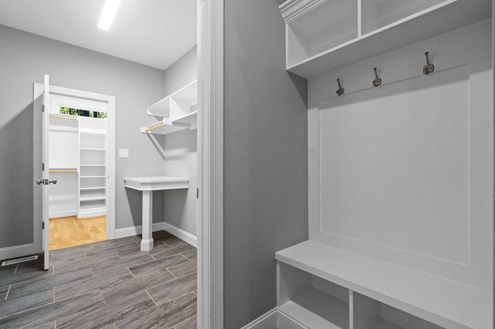 Spacious walk-in closet with gray walls and tiled floor. It features white shelving, a built-in bench with hooks, and a desk. A door opens to another bright room with more shelving and light wood flooring.