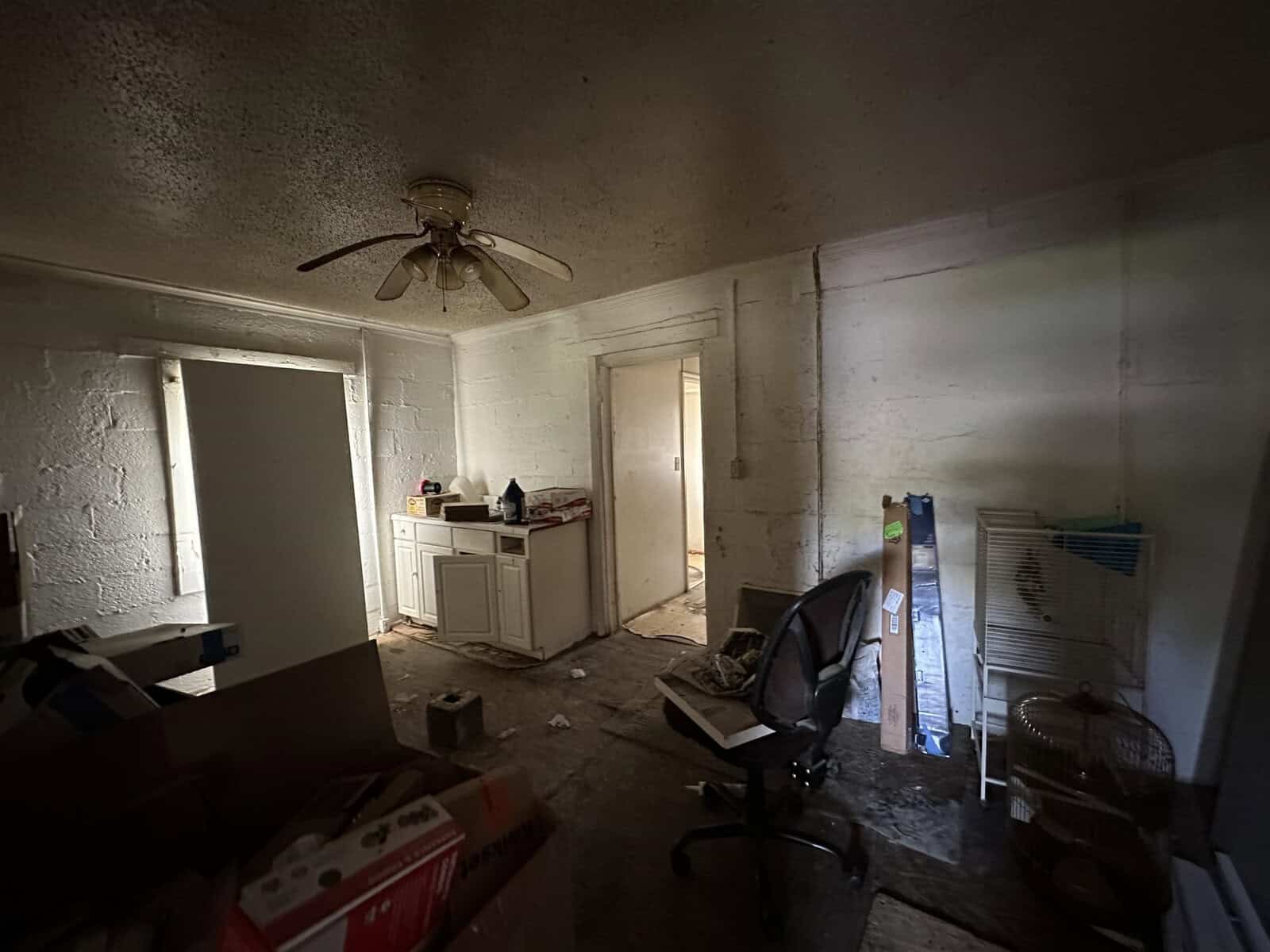 An old, dimly lit room with peeling white paint, cluttered with discarded items and furniture, featuring a ceiling fan, an open door, and signs of neglect.