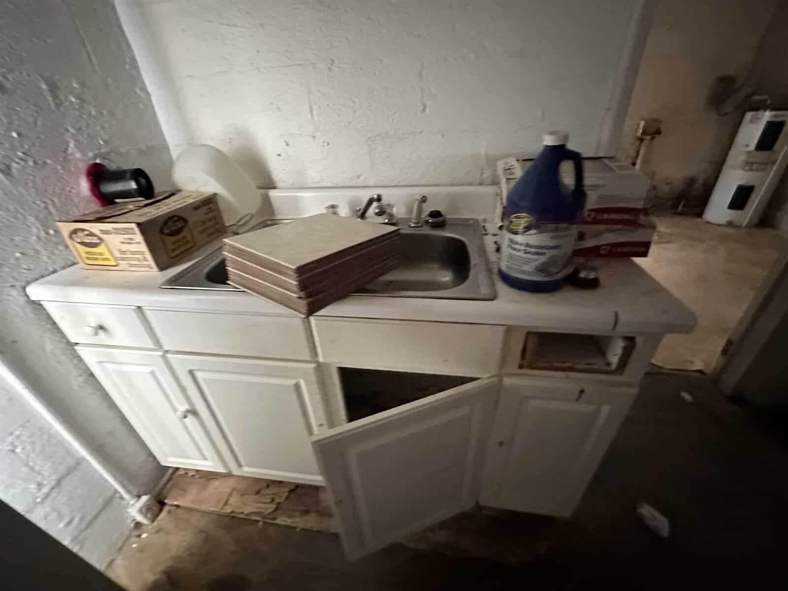 A cluttered utility sink area with cleaning supplies and stacks of tiles on a dirty, white cabinet that has one door hanging open.