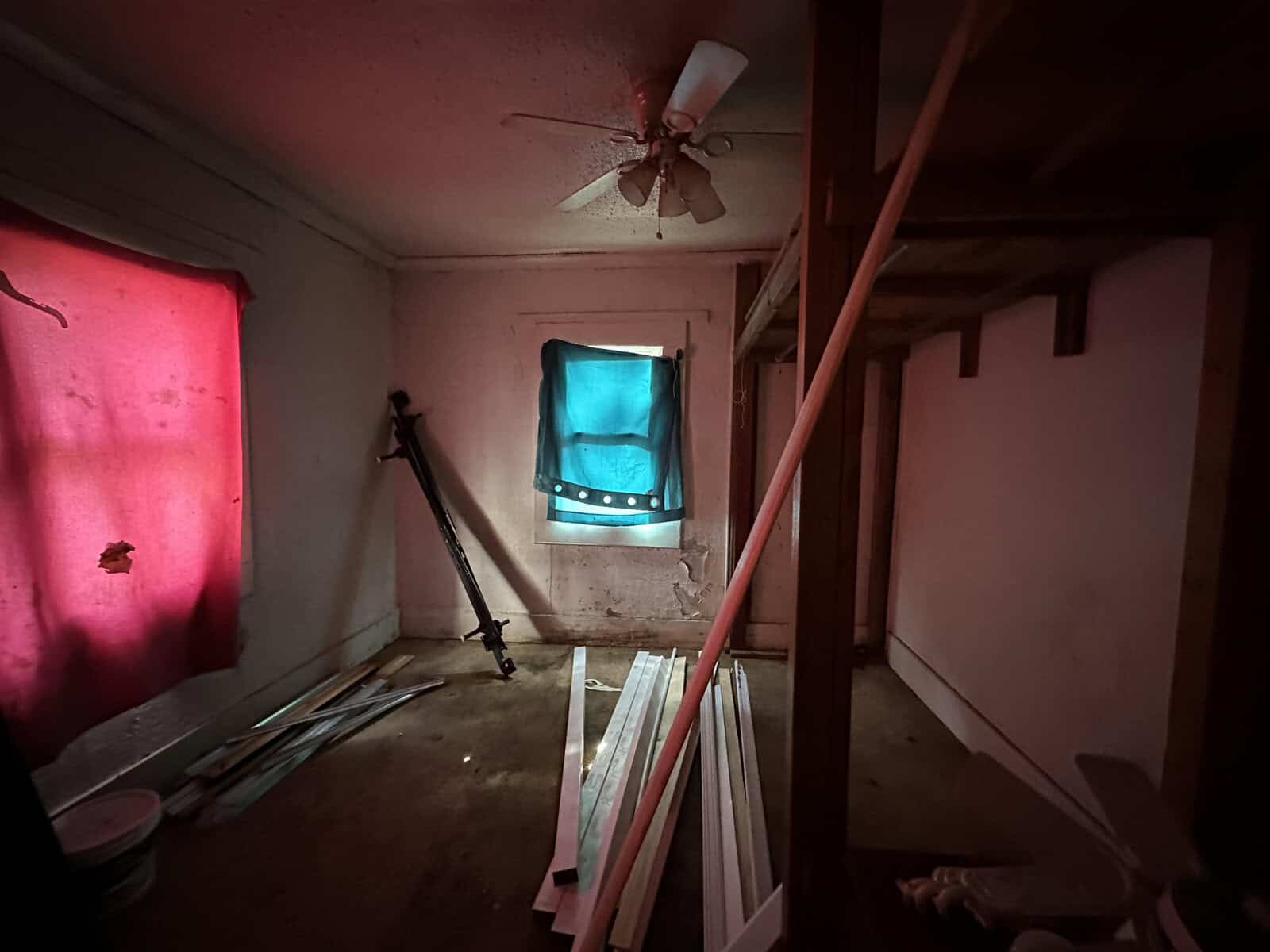 A dimly lit, neglected room with building materials scattered on the floor, a window covered by a turquoise cloth, and a ceiling fan hanging askew.