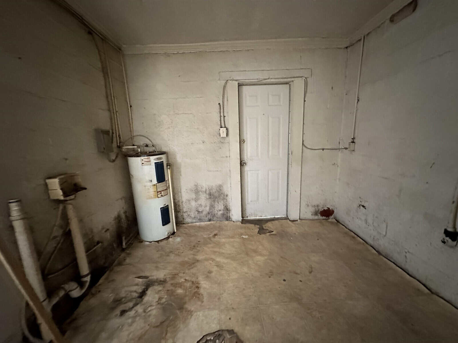 A dimly lit, neglected room with white, grimy walls and a dirty floor. a white door stands at the center, flanked by a water heater on the left and exposed pipes on the right.
