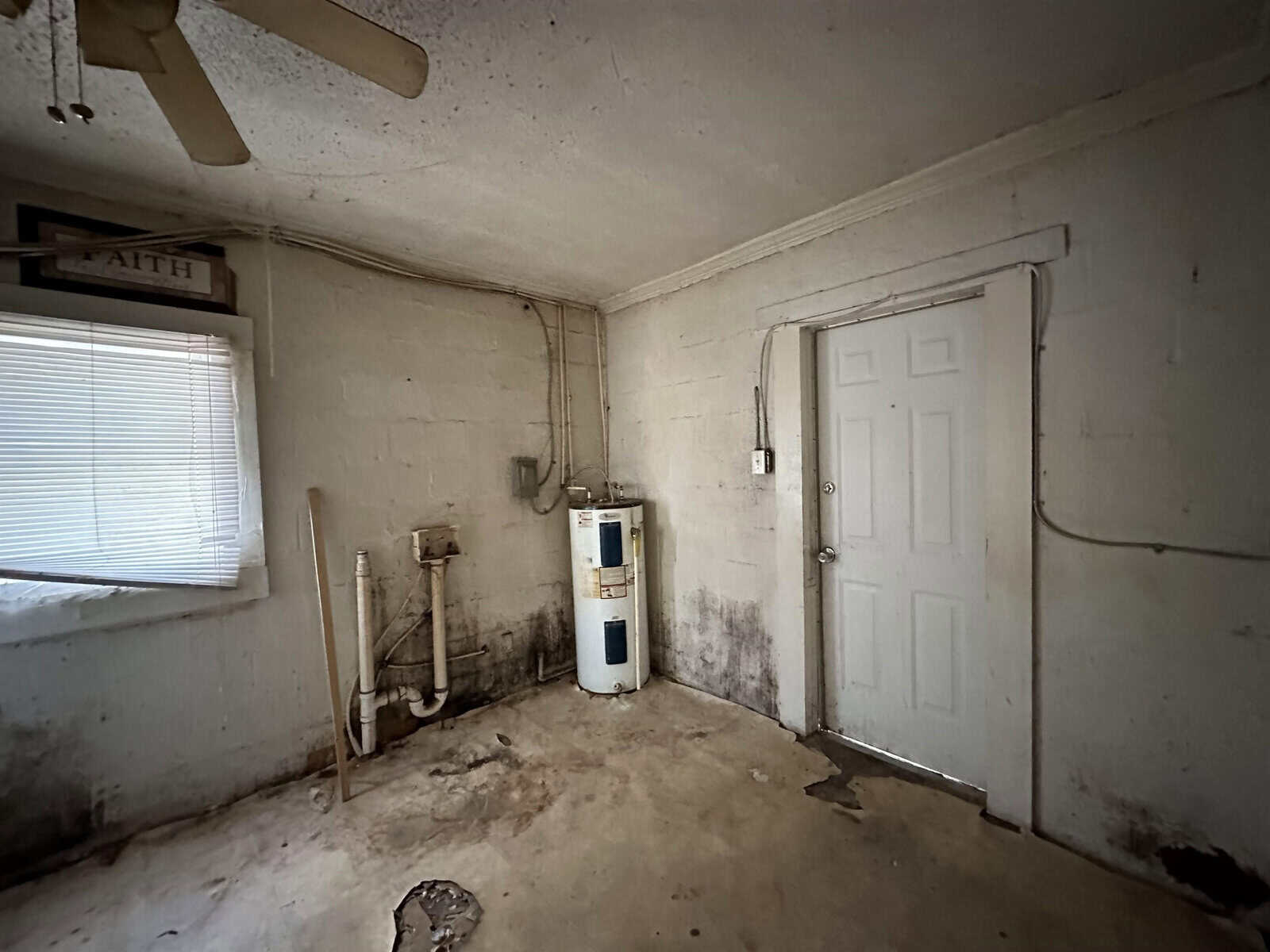 A deteriorated interior room showing signs of neglect, with a water heater to the left, a white door in the center, and a ceiling fan above, all set against walls and a floor needing maintenance.