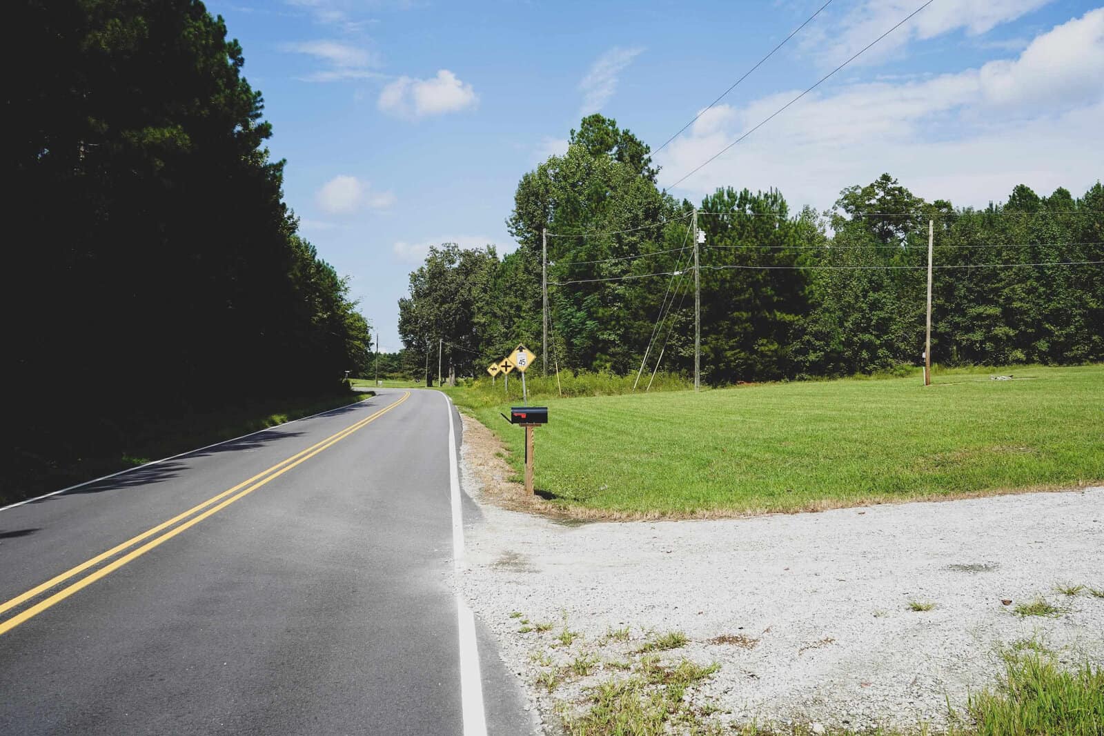 A rural road curves to the left, bordered by grass and trees. A railroad crossing sign is visible in the distance, indicating tracks ahead. A mailbox stands near the road on the right side next to a gravel-covered area.