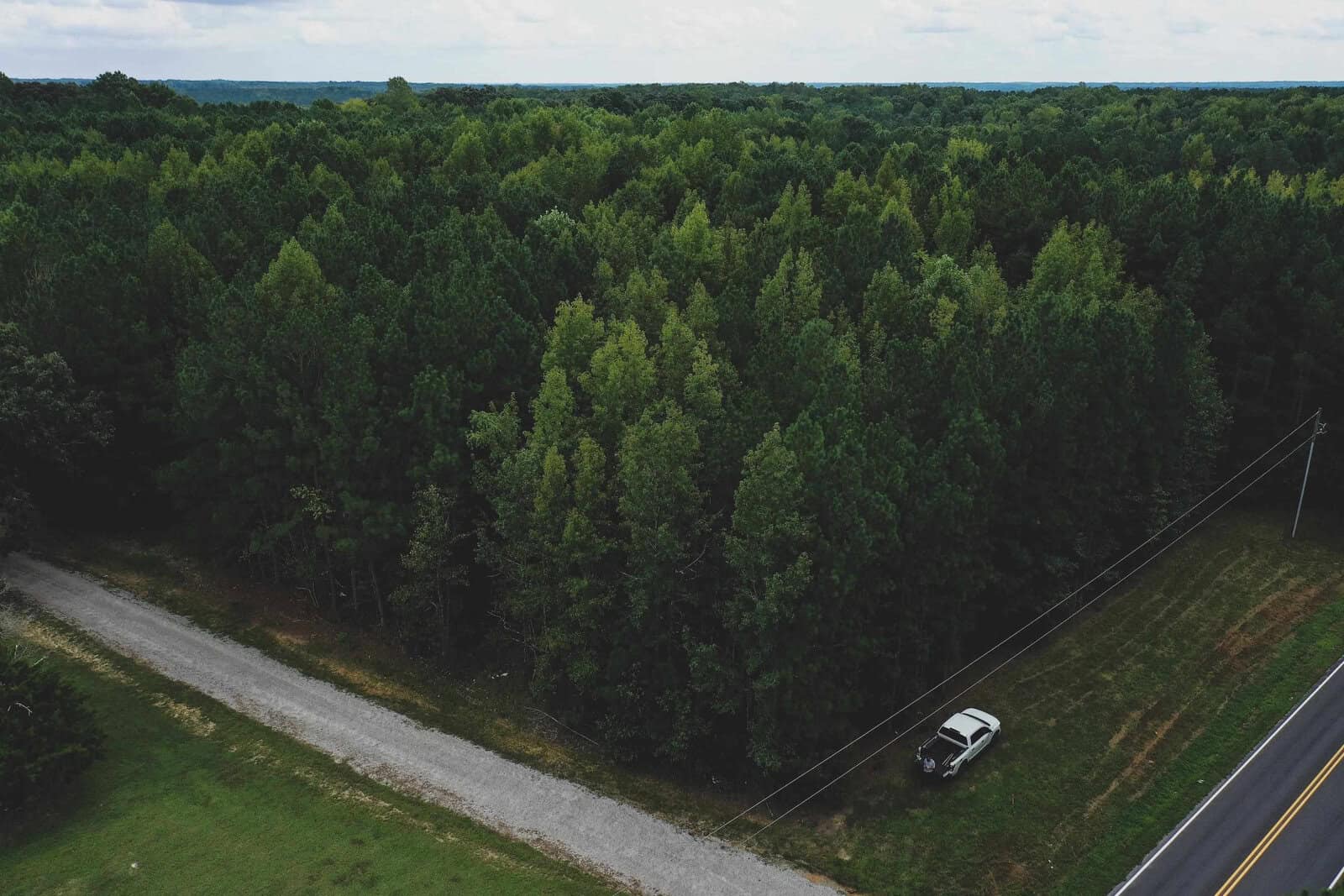 Aerial view of a lush green forest with a small dirt road on the left and a paved road on the right. A white car is parked on the grass beside the paved road. The sky is cloudy, and the horizon is lined with trees.