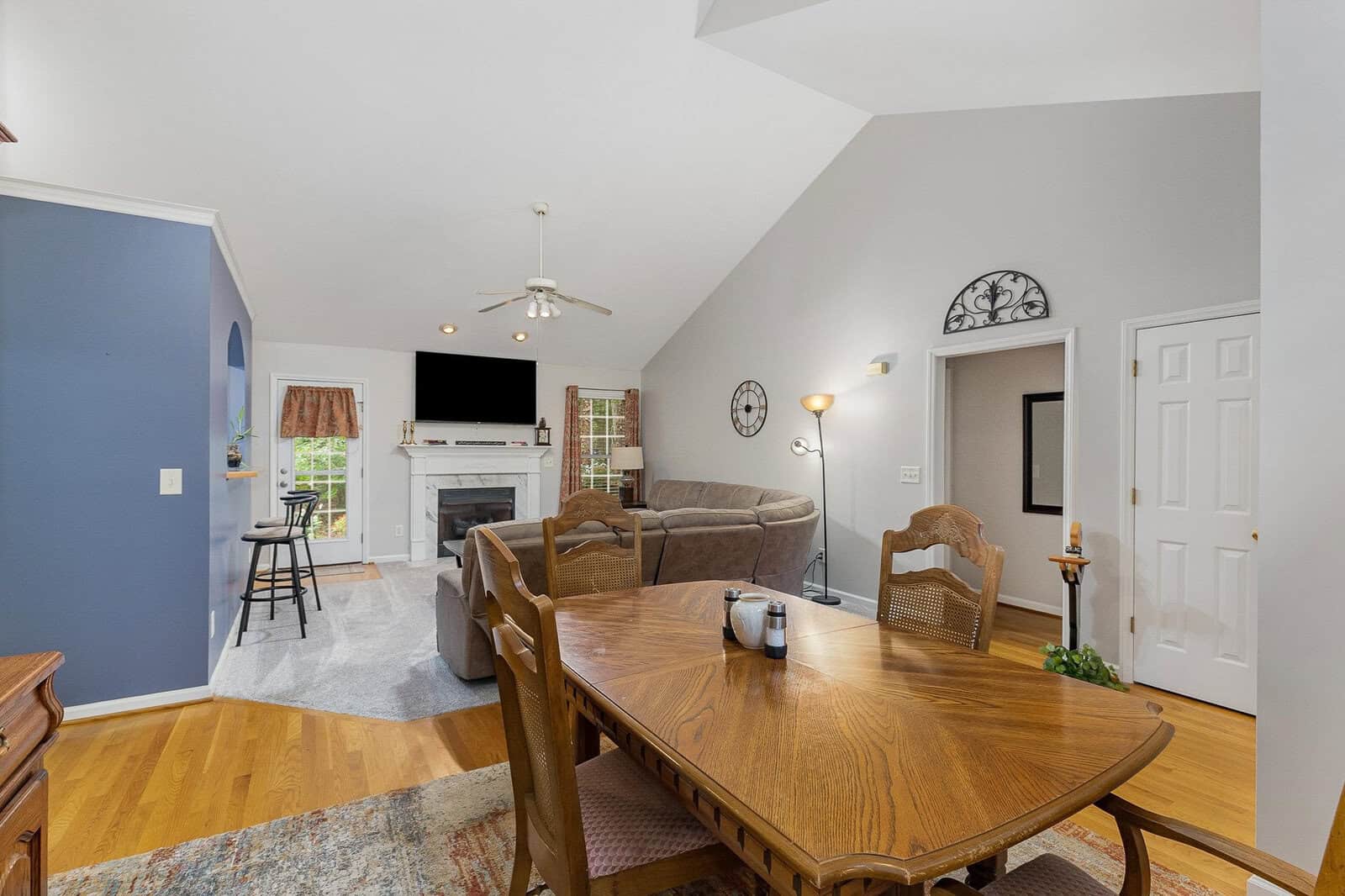 A cozy living and dining area with a vaulted ceiling. A wooden dining table with chairs is in the foreground. The living room features a sectional sofa, a fireplace, and a wall-mounted TV. Neutral walls and hardwood floors enhance the space.