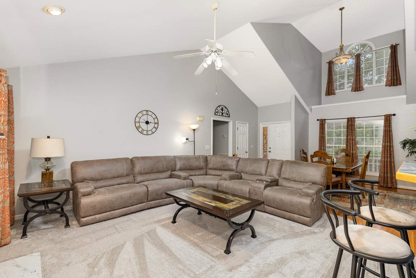 Spacious living room with beige sectional sofa, wooden coffee table, and bar stools. High ceiling with ceiling fan. Dining area in the background with large windows and curtains. Modern decor with a wall clock and carpeted floor.