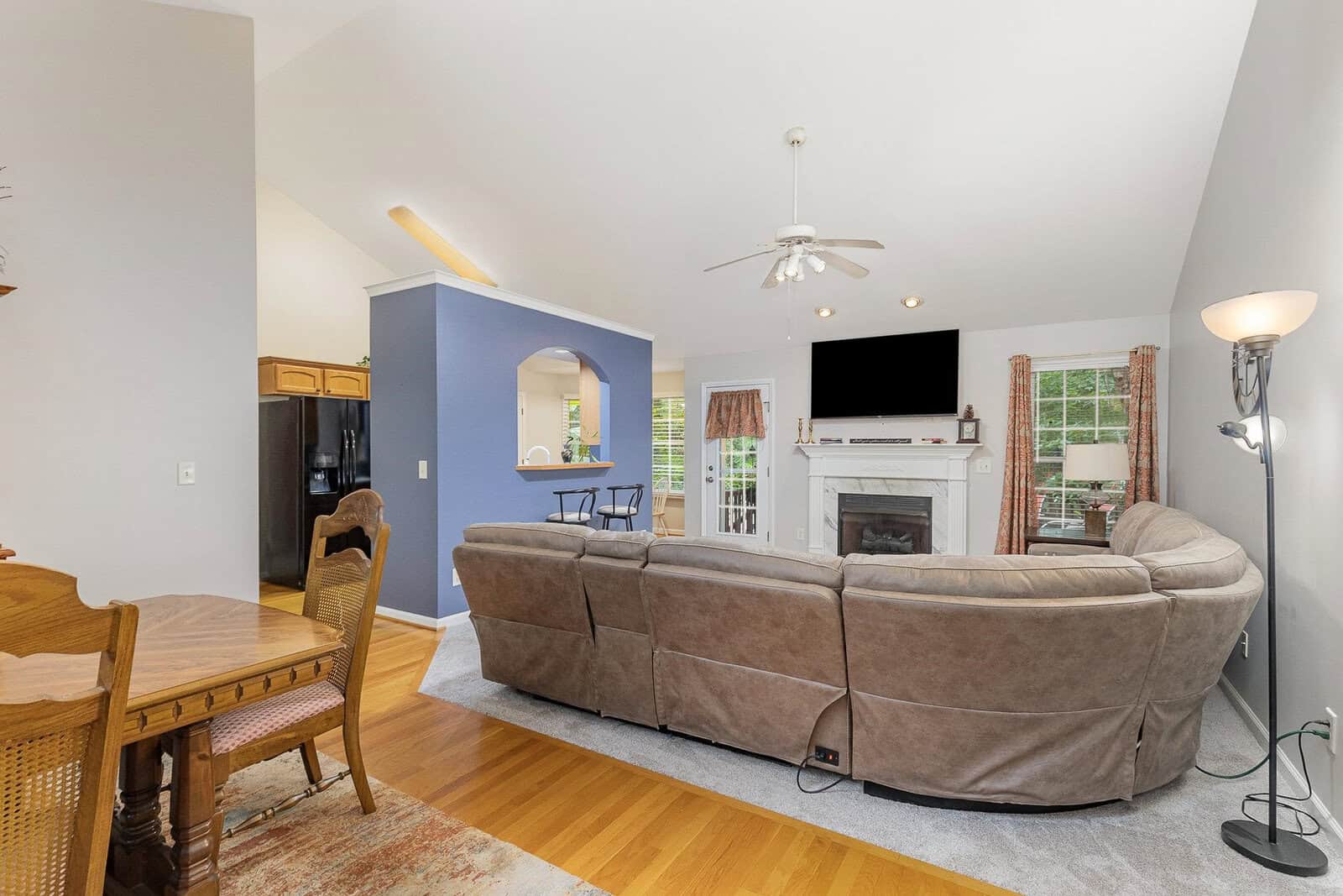A living room with a large beige sectional sofa facing a fireplace and wall-mounted TV. The room has hardwood floors, a high ceiling with a fan, and a window with curtains. A dining table and chairs are visible to the left. The kitchen is in the background.