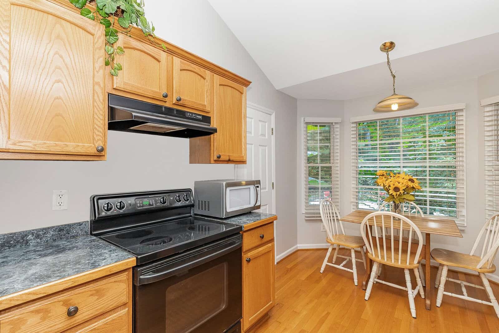 A kitchen with wooden cabinets, black countertops, and a black stove. A small dining table with four chairs is set by a window with blinds, adorned with a vase of sunflowers. A microwave sits on the counter, and a hanging light fixture is above the table.
