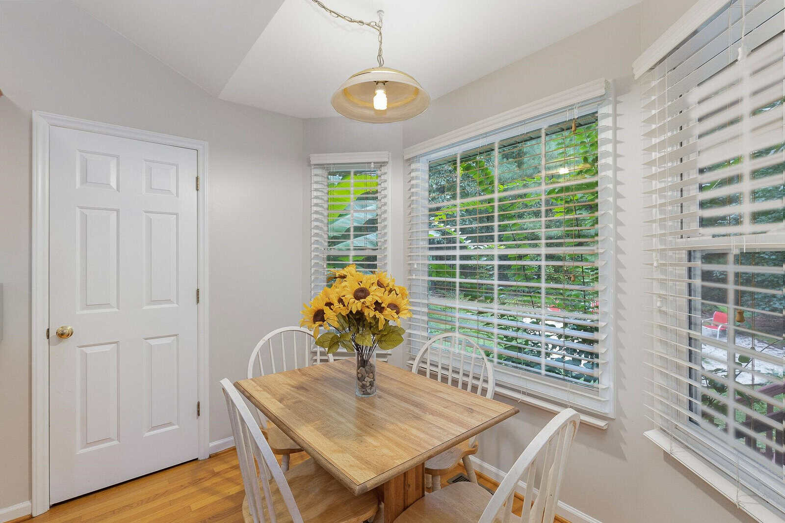 A cozy dining nook with a wooden table and four white chairs. A vase with sunflowers sits on the table. Large windows with blinds overlook a green, leafy garden. A pendant light hangs above the table, illuminating the space.