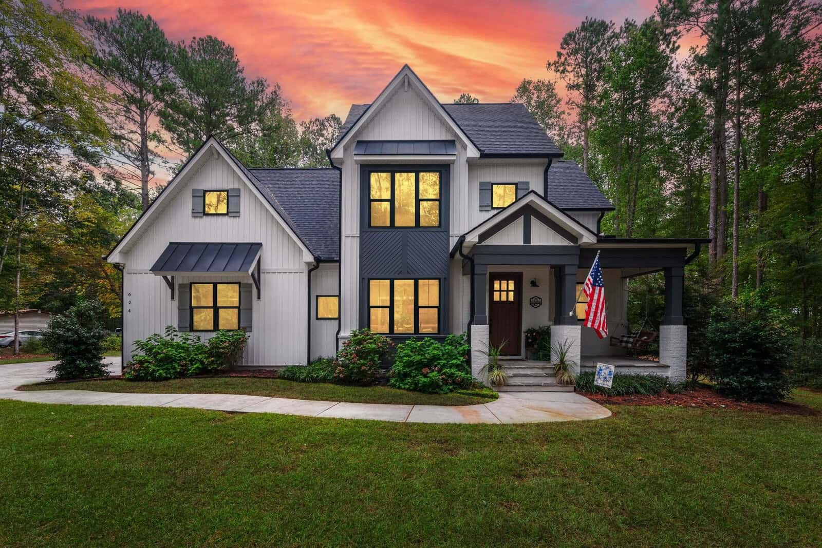 A modern two-story house with a white and dark gray exterior, surrounded by lush greenery. The front porch has an American flag. The sky is vibrant with a pink and orange sunset. A paved walkway leads to the entrance.