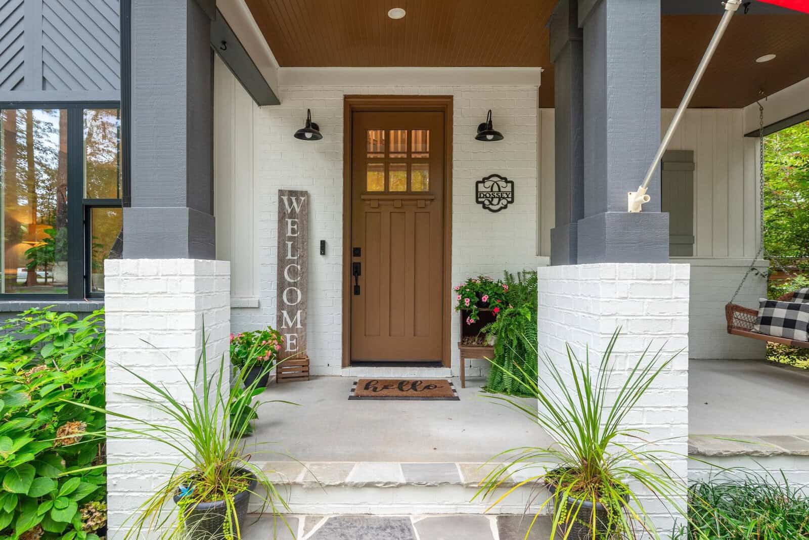 A cozy porch entrance with a brown door and "Welcome" sign. Potted plants adorn the steps, and a doormat reads "hello." Black sconces light the white brick walls. A hanging swing is partially visible on the right.