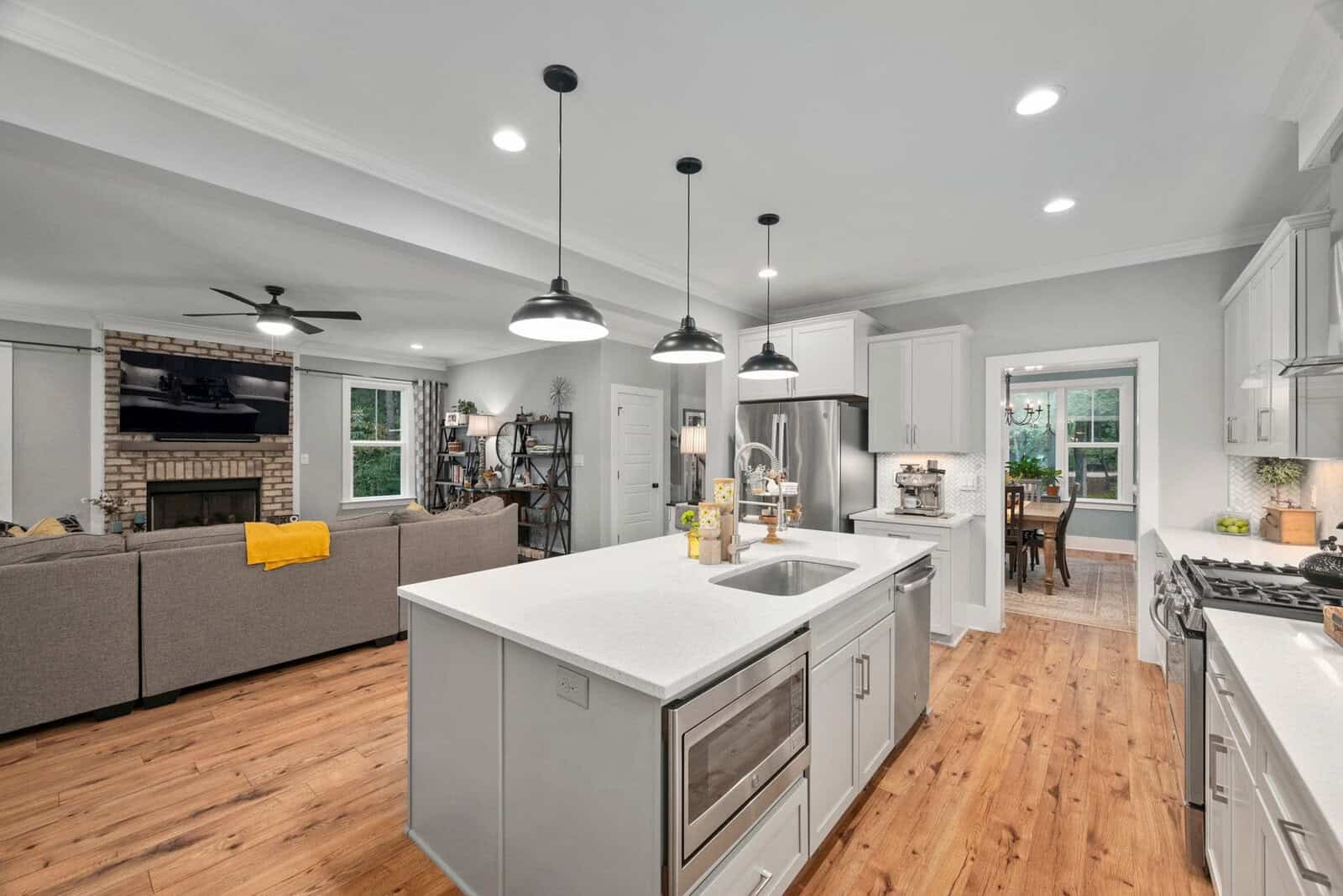 Modern open-concept kitchen and living room with a large white island, stainless steel appliances, and wood flooring. The living area features a gray sofa, brick fireplace, and ceiling fan. Pendant lights hang above the island, and a dining room is visible.