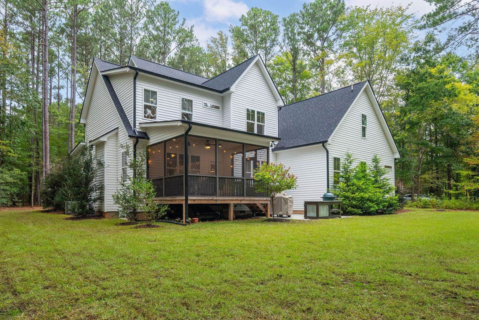 A white two-story house with a dark roof is surrounded by green trees. It features a screened porch with large windows and a neatly maintained lawn in the foreground, on a clear day.