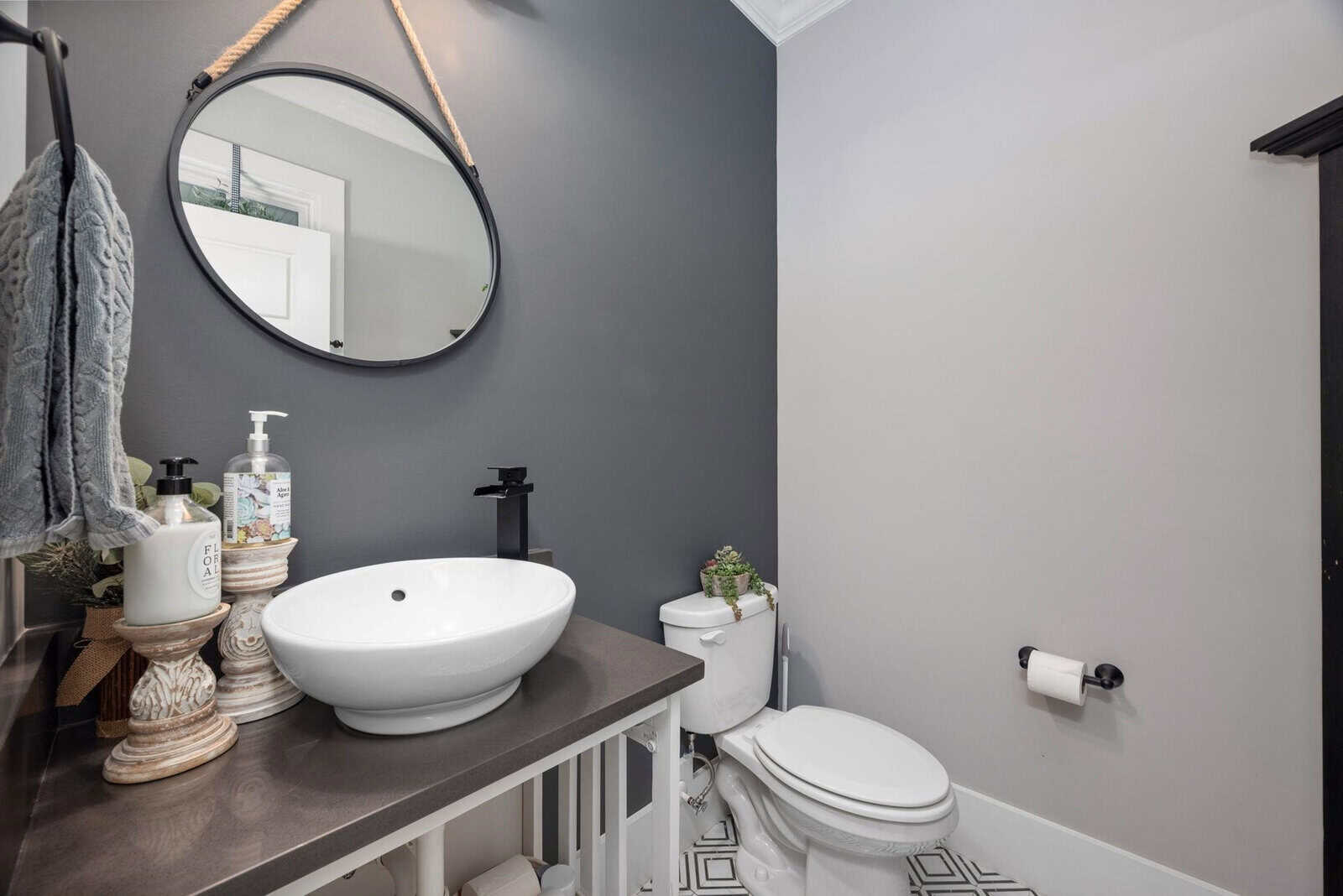 A modern bathroom with a round mirror above a black countertop with a white vessel sink. There's a potted plant, soap dispenser, and towels on the counter. A toilet with a roll of toilet paper is beside it. Walls are gray, and the floor is patterned.