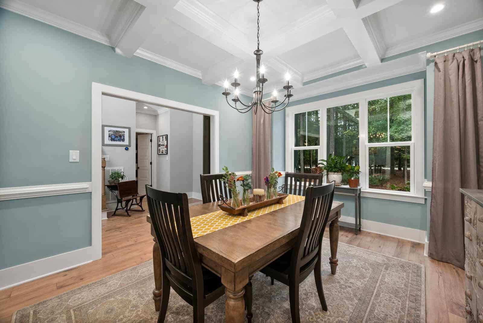 A dining room with a wooden table set for four, featuring a runner, flowers, and candles. Black chairs surround the table. There's a chandelier overhead, and large windows overlook a garden. The walls are light turquoise, and hardwood floors are present.