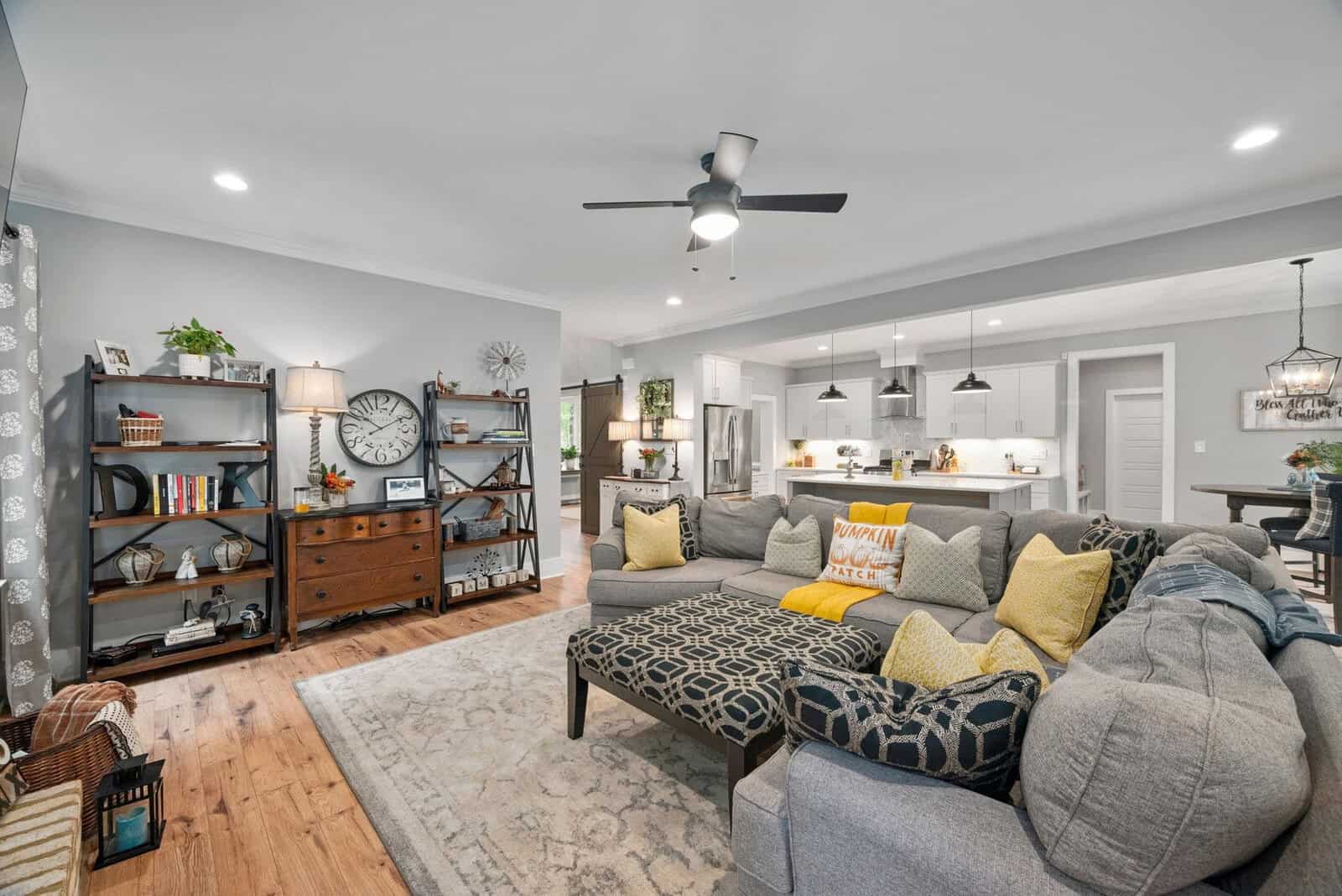 A cozy living room with a gray sectional sofa adorned with yellow and patterned pillows. There's a rug on wooden flooring, a ceiling fan, shelves with books and decor, and an open view of a kitchen with white cabinets and pendant lights.