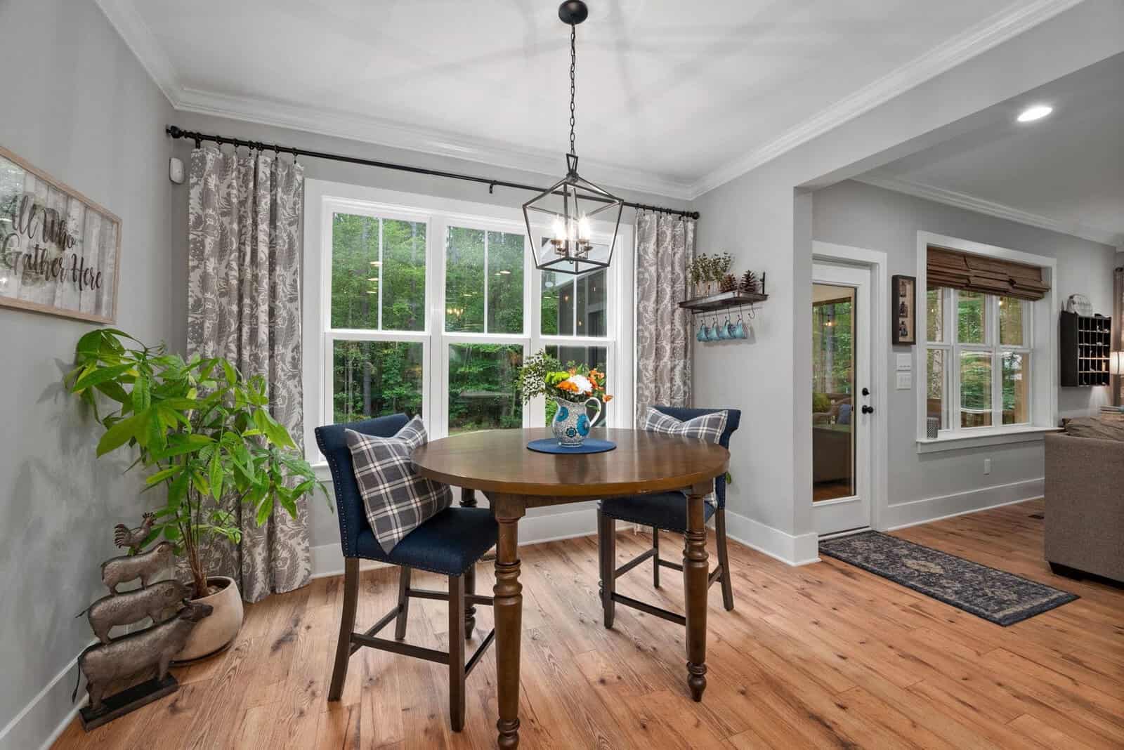 A cozy dining area features a round wooden table with two plaid-cushioned chairs. A vase with flowers sits on the table. Large windows with patterned curtains provide a view of greenery. Soft lighting and wooden floors add warmth to the room.