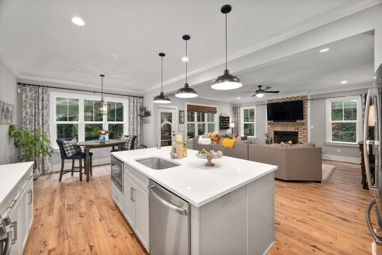 Modern open-concept kitchen and living area. Features a white kitchen island with a sink, pendant lights, and hardwood floors. Cozy living room with a brick fireplace and a dining nook with large windows and plants. Neutral decor and ample lighting.