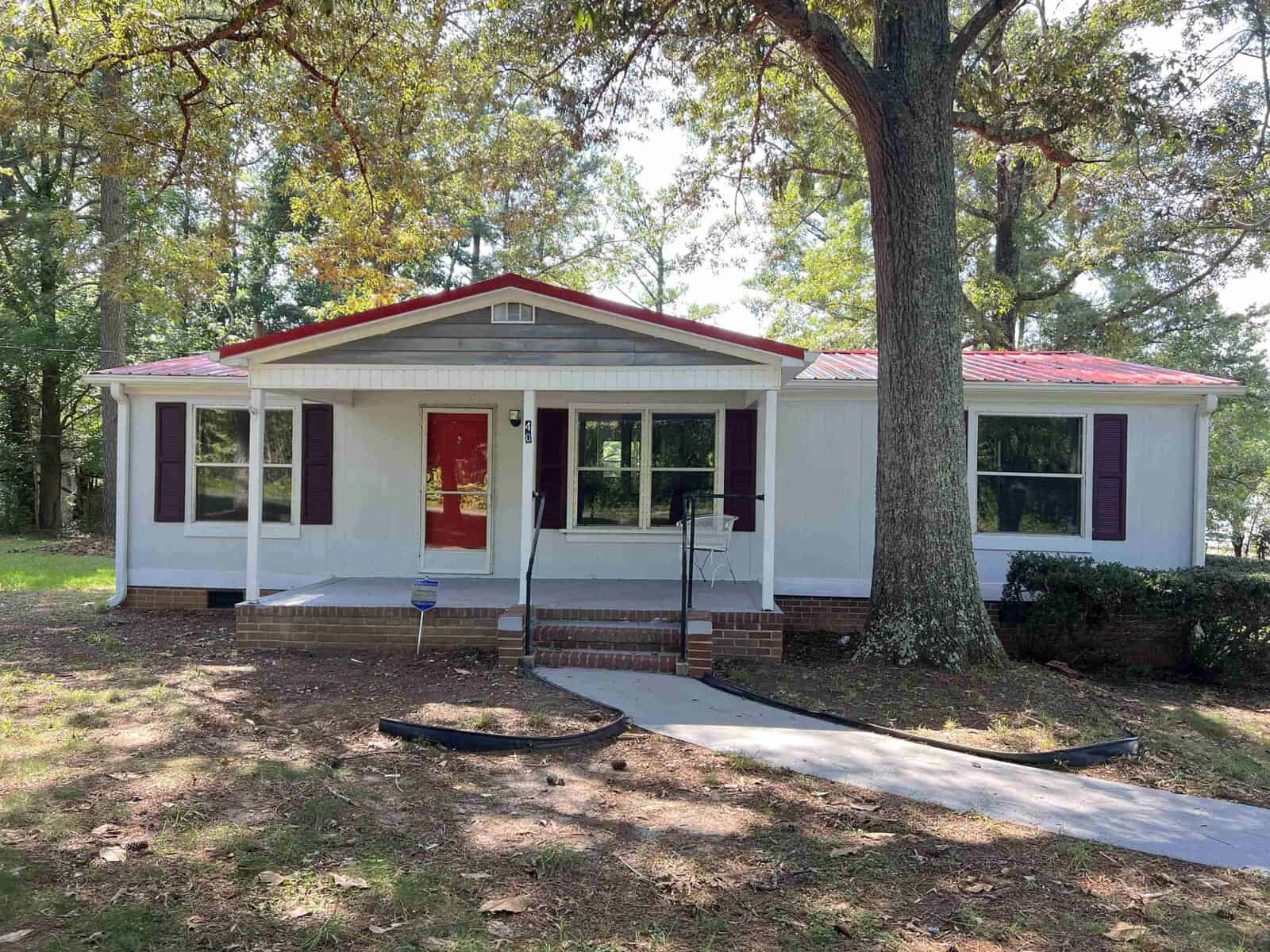 A single-story house with white siding, red roof and door, and a front yard with a large tree on a sunny day.