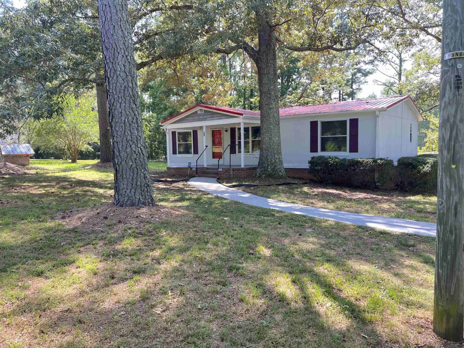 A single-story house with a white and purple facade, a red door, and a front yard with trees under a clear sky.