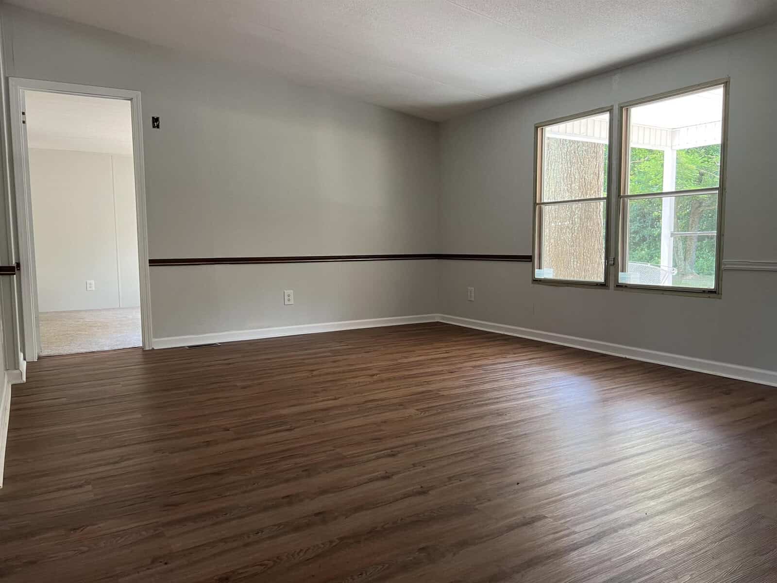 An empty room with light gray walls, a dark trim near the floor, hardwood flooring, and a large window on the right. an open doorway is seen on the left side leading to another room.