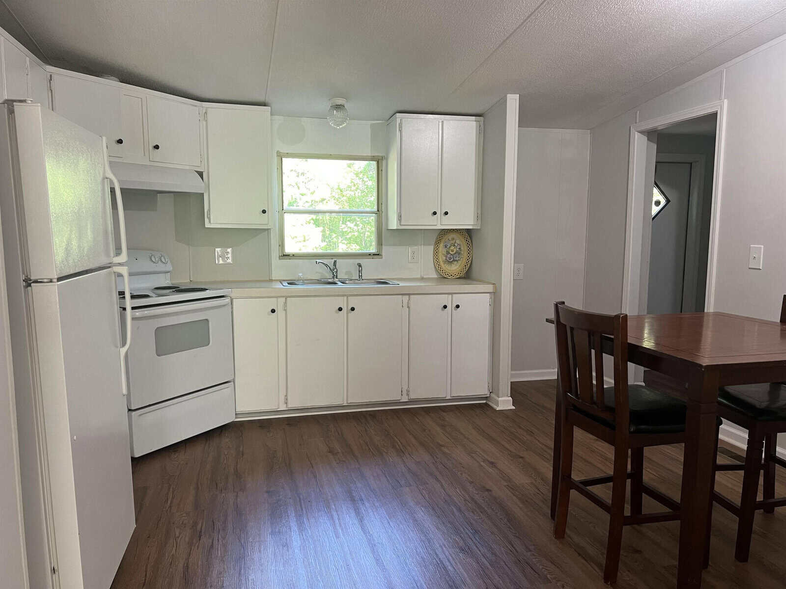 A modest kitchen interior with white cabinetry and appliances, featuring a wooden dining table and chairs, with a view of two windows allowing natural light.