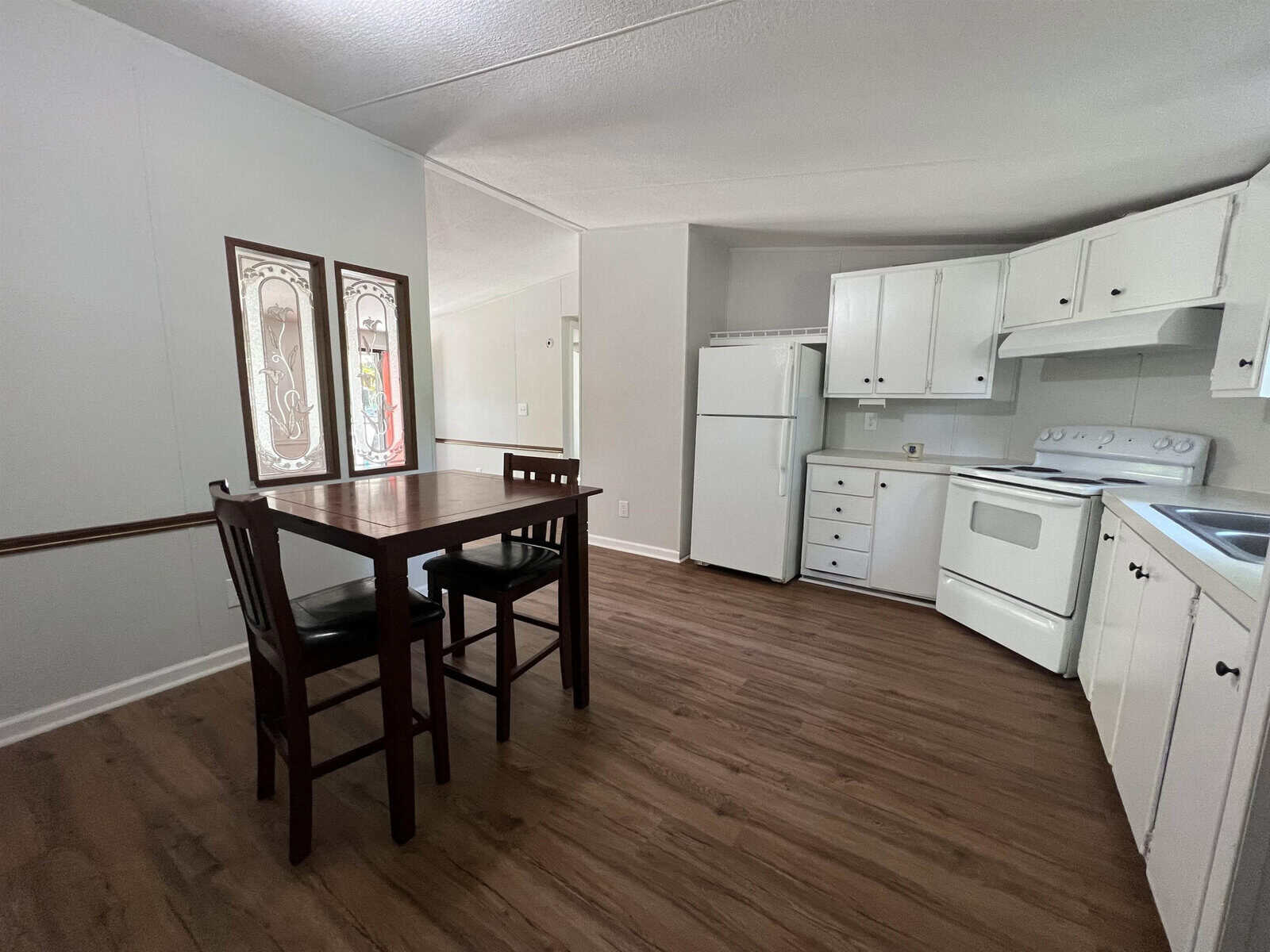 A sparsely furnished kitchen and dining area with wooden flooring, a wooden table with one chair, white appliances, and white cabinetry, accentuated by a decorative painting on the wall.