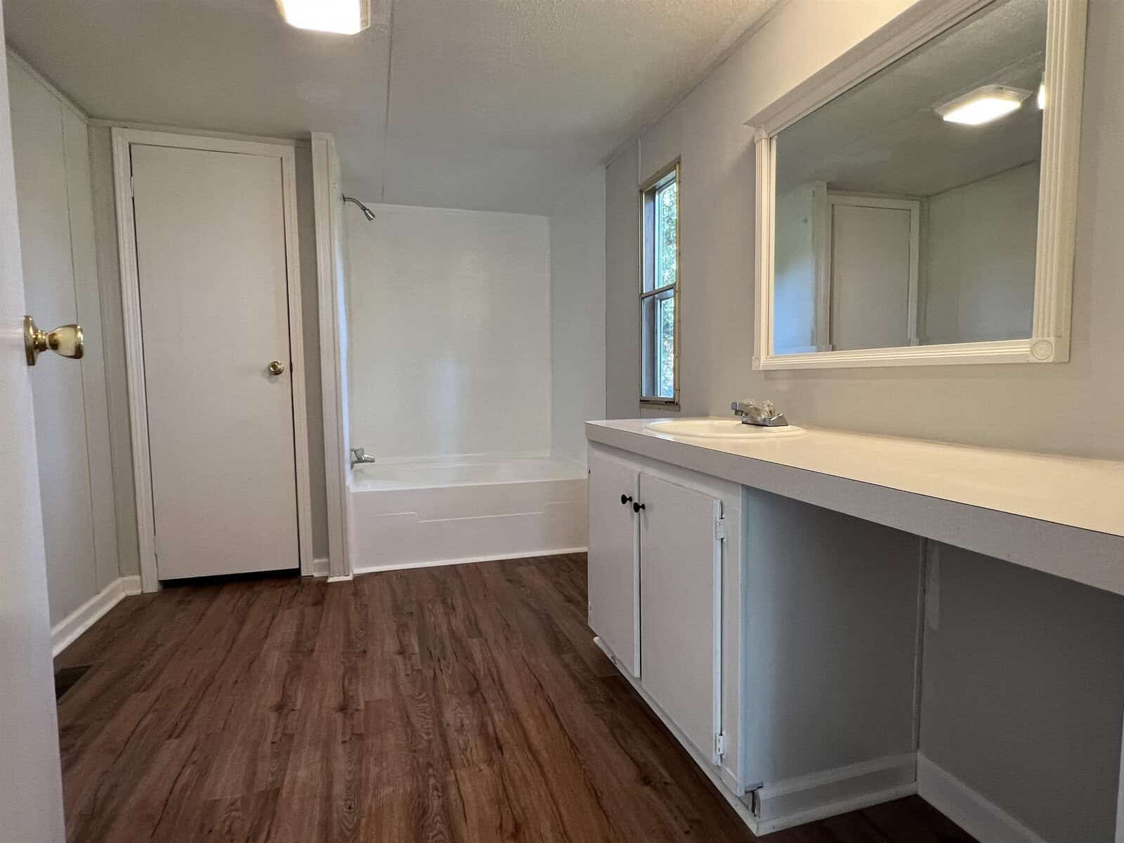 An empty room featuring laminate flooring, a built-in countertop with cabinets, and a bathroom visible through an open doorway on the left. a window with blinds is on the right, and there is a mirror above the countertop.
