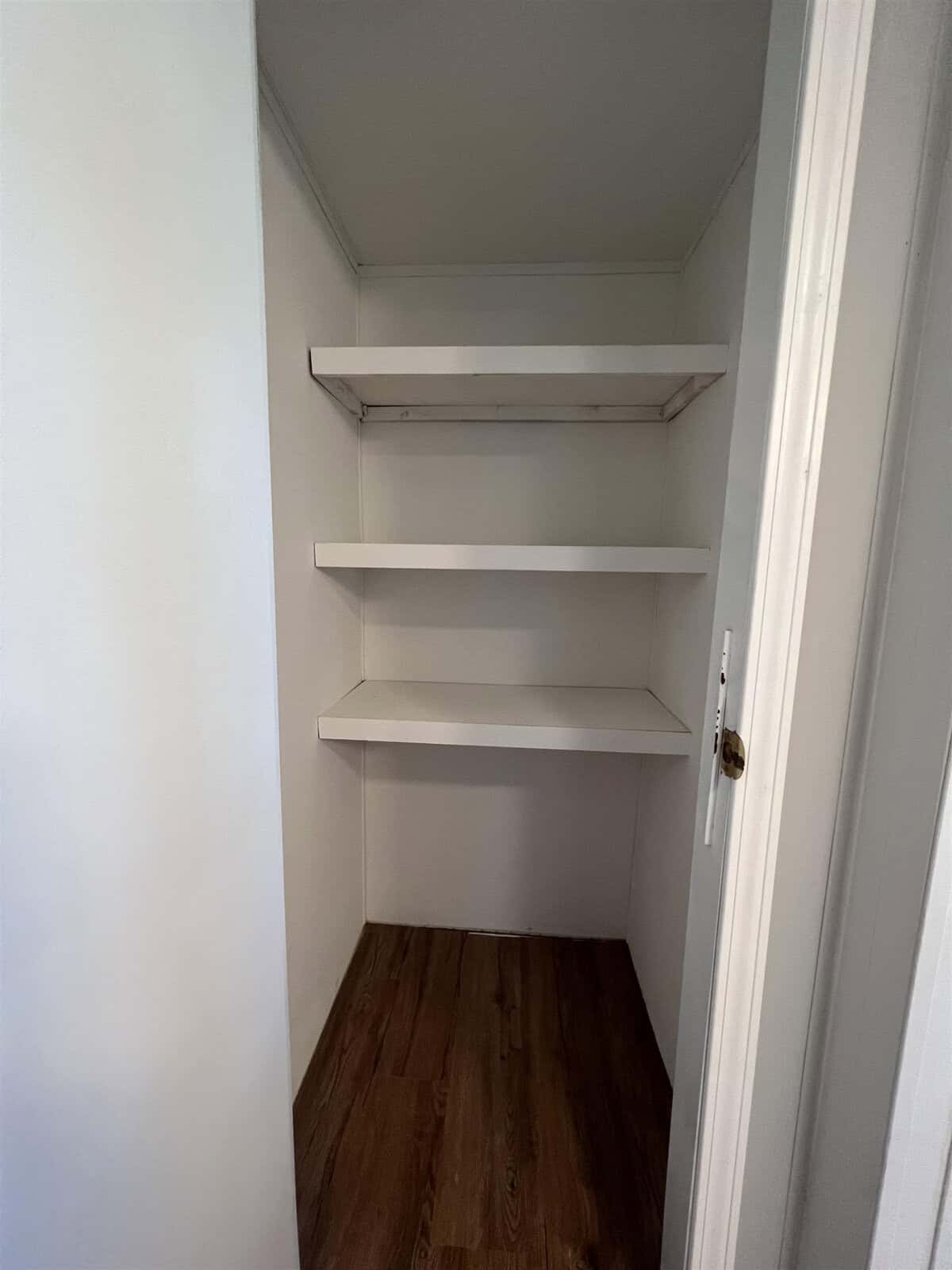 An empty closet with three white shelves on a wall and wooden flooring.