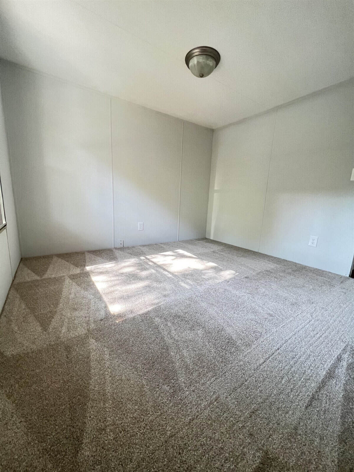 An empty room with beige carpeting, white walls, and natural light coming in from the windows, casting shadows on the floor.