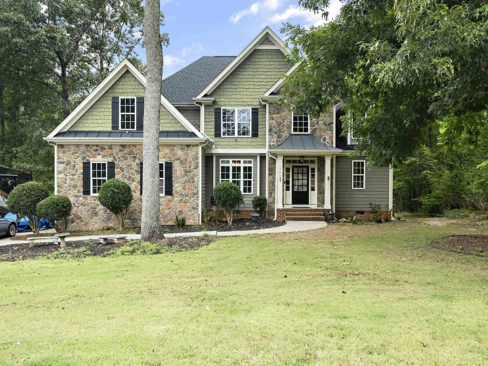 A large, two-story house at 1703 Creekview Drive in Franklinton boasts a stone and green siding exterior. It features multiple windows and a black front door. The well-maintained lawn, adorned with small shrubs and trees, is set against a backdrop of tall trees.